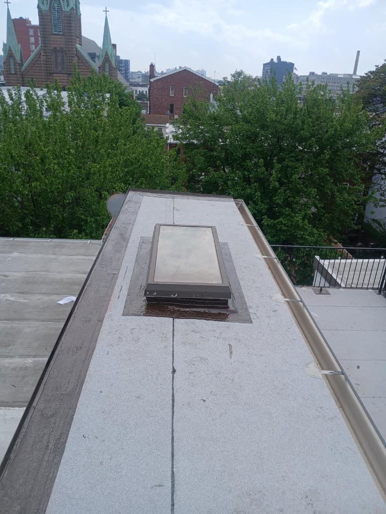 Narrow roof section with new gray granulated cap sheet, a skylight on a curb flashed in black, a brown-coated wall edge on the left and a tan metal gutter along the right edge