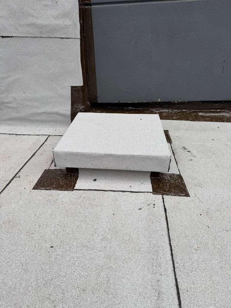 Small raised box wrapped in gray granulated cap sheet on brown-sealed corner pads, set on a flat roof beside a wall flashed in brown