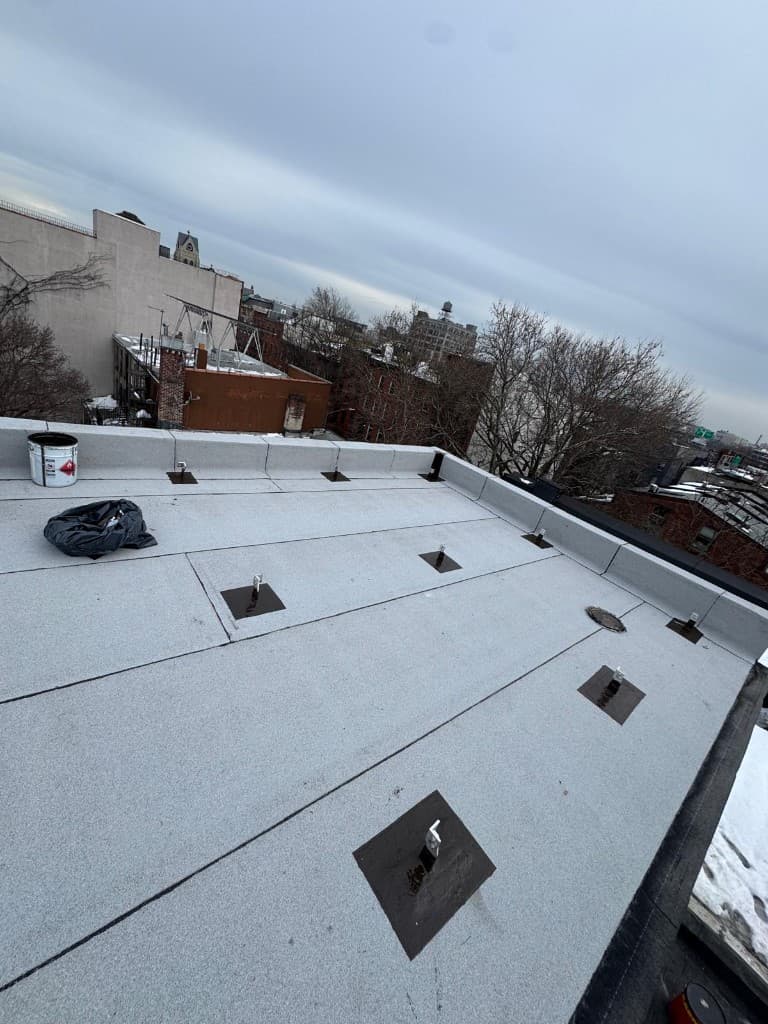 Finished bulkhead roof with new gray granulated cap sheet and visible lap lines, parapets flashed in the same sheet, metal anchor posts on black flashed pads, a round roof drain and a paint pail on the parapet