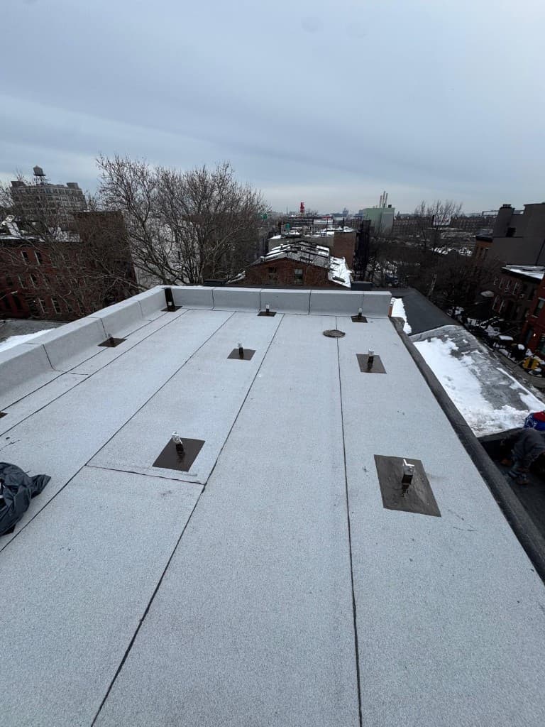Wide view of a finished bulkhead roof with new gray cap sheet, flashed parapets on two sides, anchor posts on black pads and a roof drain, with city rooftops behind