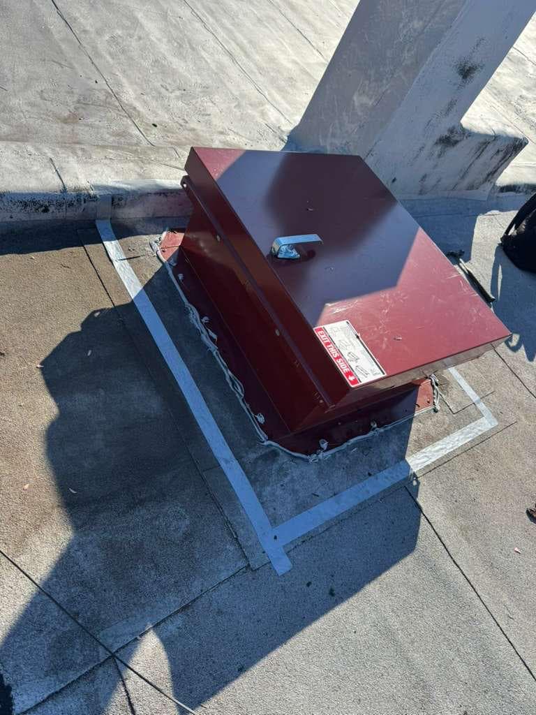 Maroon roof hatch with a handle and an exit label on its curb, with a sealant bead along the base and a white painted outline on the roof around it