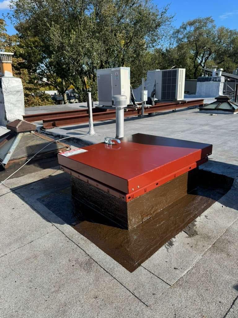 Red metal roof hatch with a handle and warning label on a curb coated in glossy brown, with the brown coating carried out onto the gray roof around it, a vent pipe and skylights beside it