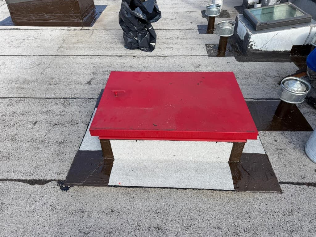 Red roof hatch on a curb wrapped in gray granulated cap sheet with brown-sealed corners, and vent pipes with metal caps set in glossy brown pads beside it