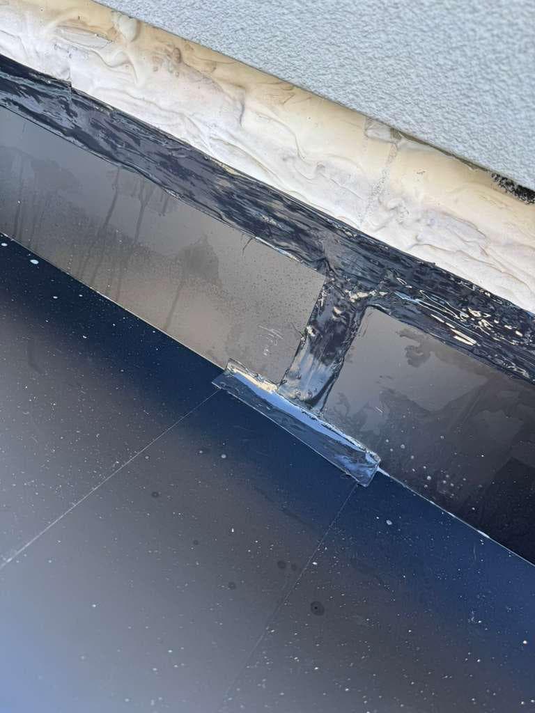 Close view of glossy black membrane flashing turned up a gray wall, with a reinforcing patch over a seam and a white sealant bead along the top edge