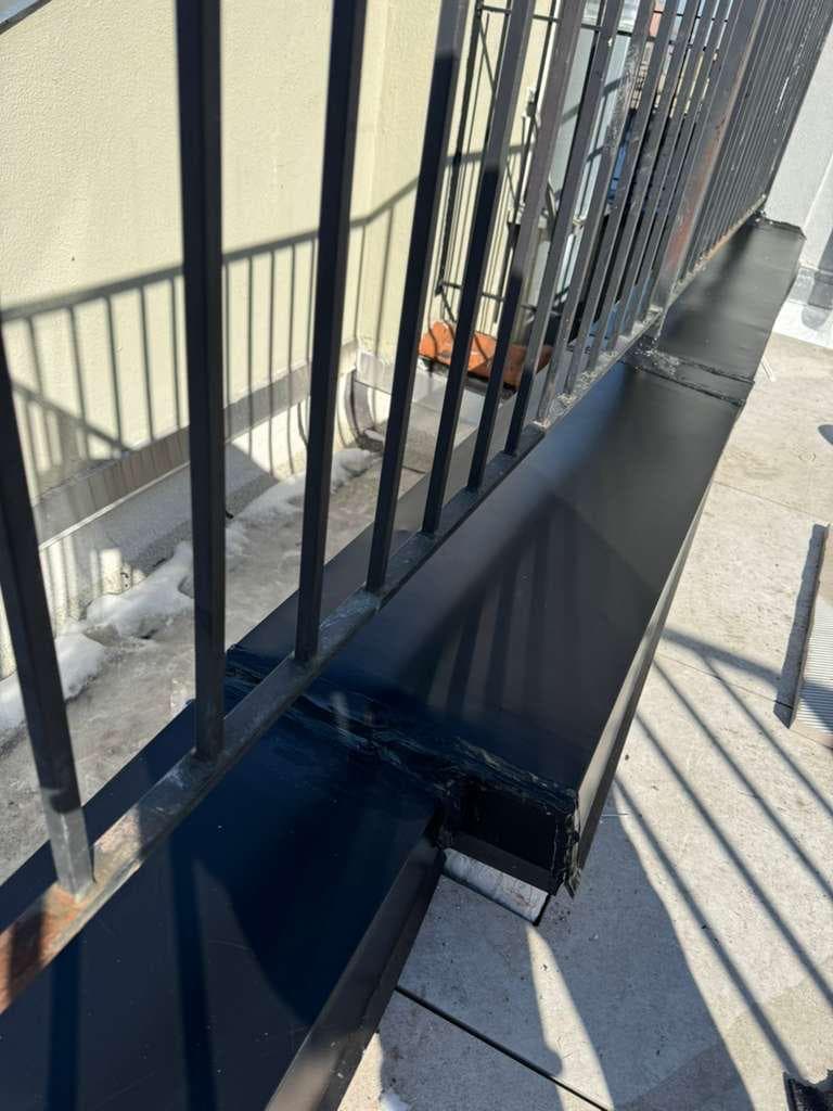 Black sheet-metal cap covering a low roof wall, formed around the posts of a metal railing, with sealed joints and a folded end