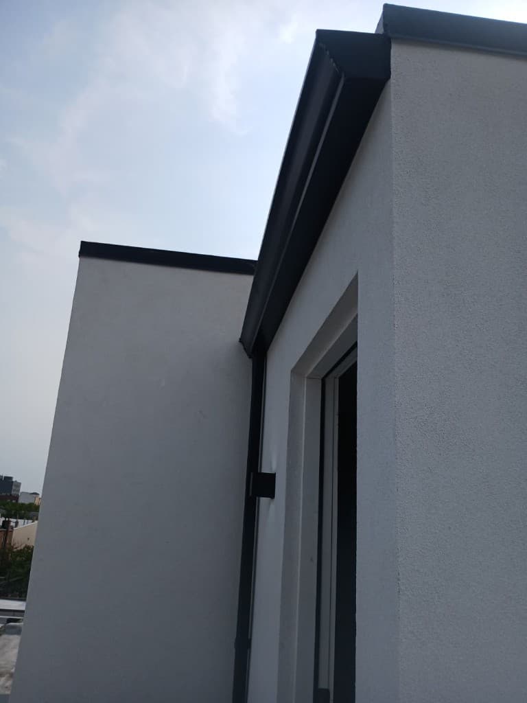 Black metal gutter sloping along the roofline of a white stucco wall, with a black downspout run down the wall corner