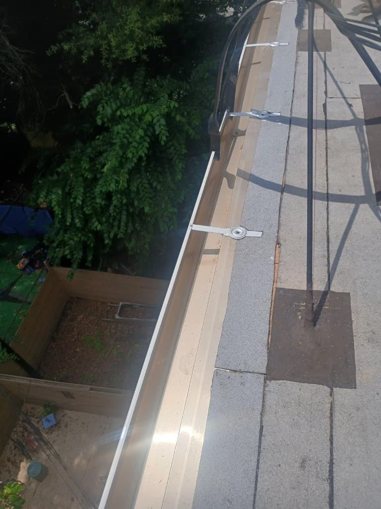 Metal gutter running along a roof edge, held with flat strap hangers fastened into the roof, with granulated roofing membrane alongside