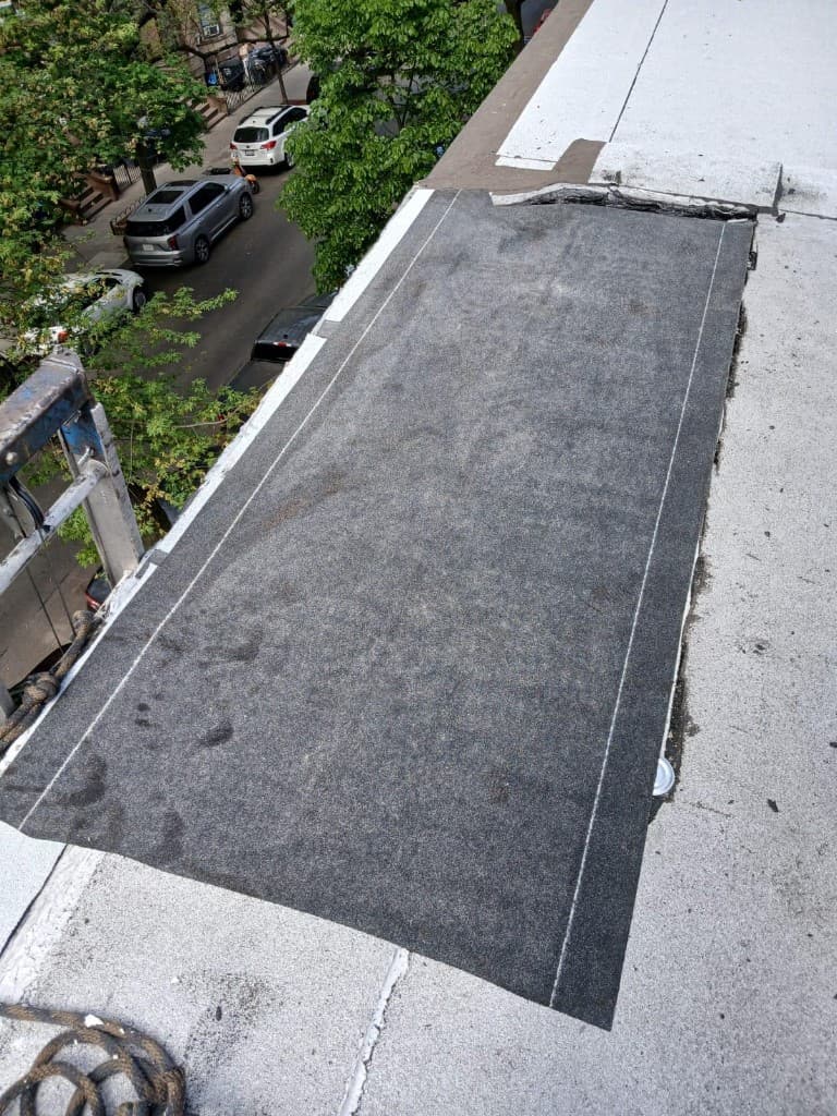 Strip of new dark base sheet laid along a roof parapet with chalk lines on it, a ladder against the edge and cars on the street below