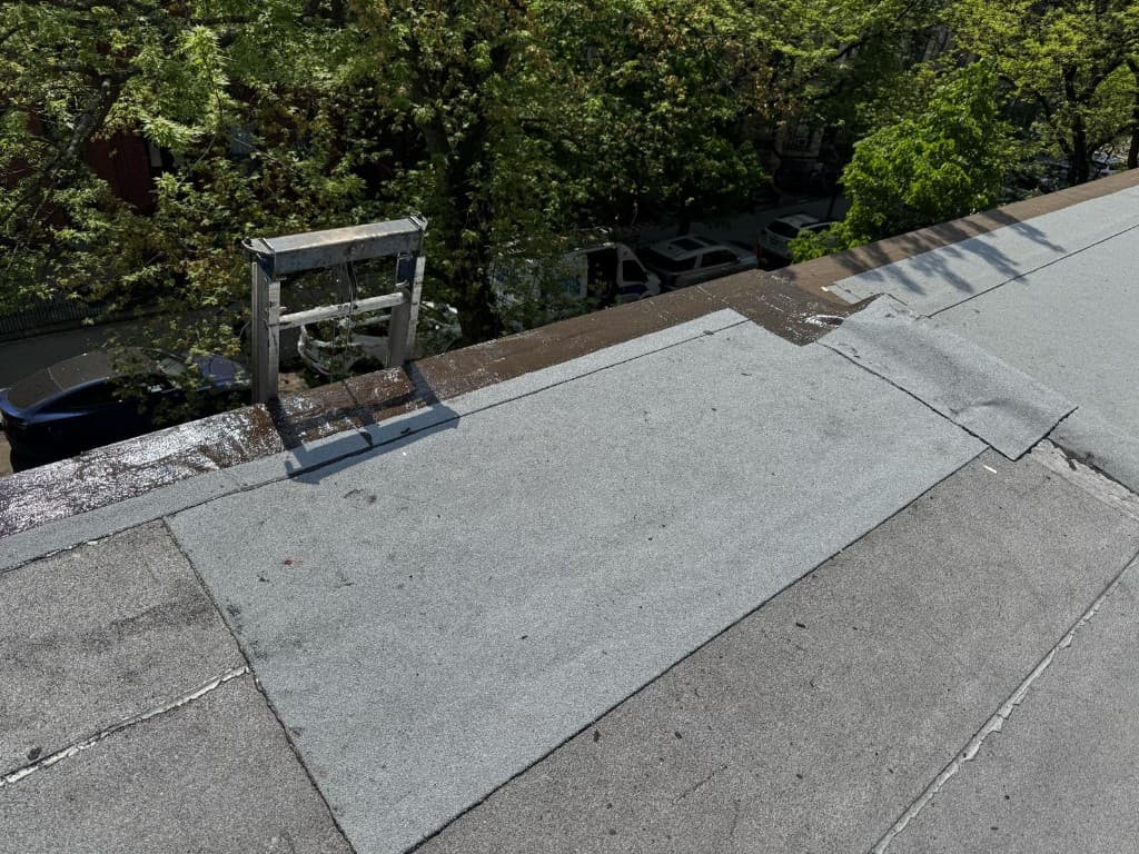 Corner of a flat roof repair where new gray granulated cap sheet laps over the existing roof beside a parapet edge sealed in brown, with a ladder at the edge