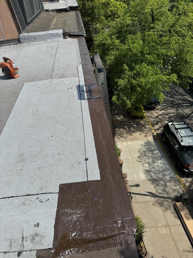 Section of new gray granulated cap sheet beside a parapet whose top and corner are coated in brown, with a ladder and a leaf blower on the roof