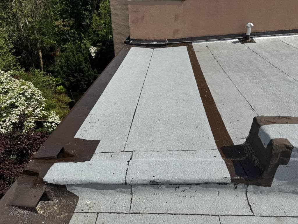 Strips of new gray granulated cap sheet lapped into a flat roof, with the parapet edge and corners sealed in brown and a vent pipe at the far wall