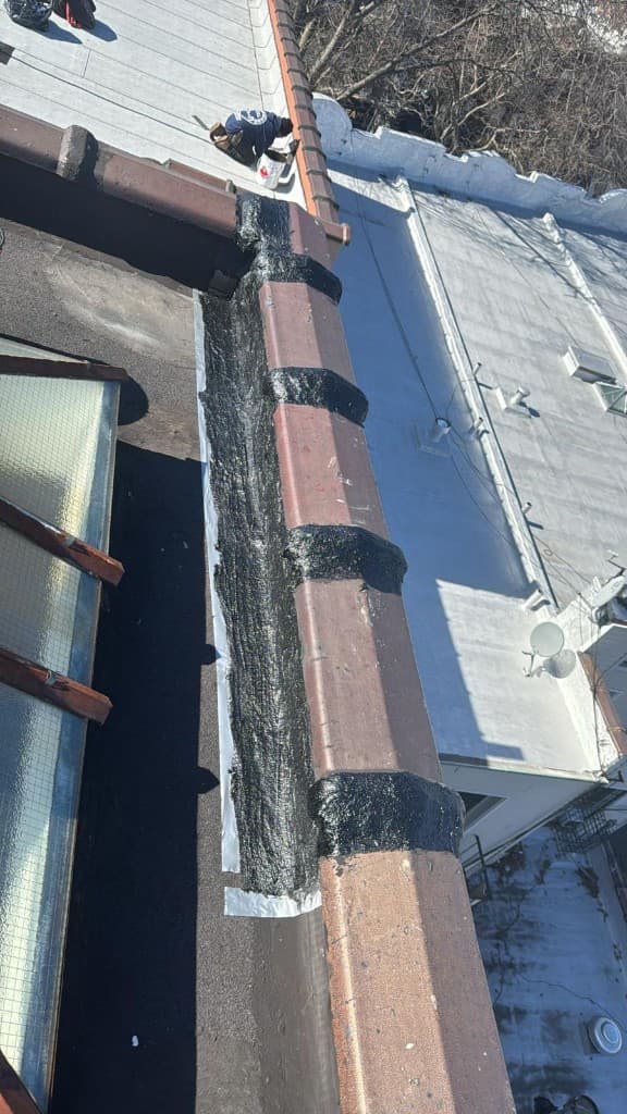 Clay coping tiles along a parapet with the joints between the tiles sealed in black, a skylight on the left and a roofer working at the top of the photo