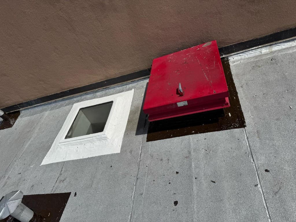 Red roof hatch and a skylight with a white-coated curb on a gray roof, a vent pipe flashed in black, and the base of a tan stucco wall flashed in black with a white finish
