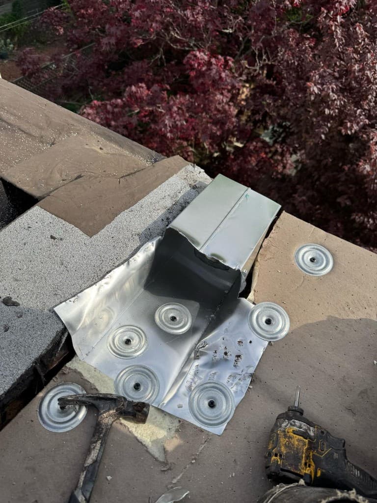 Galvanized sheet-metal outlet set through a parapet and fastened to new roof boards with round plates, with a hammer and a drill on the roof