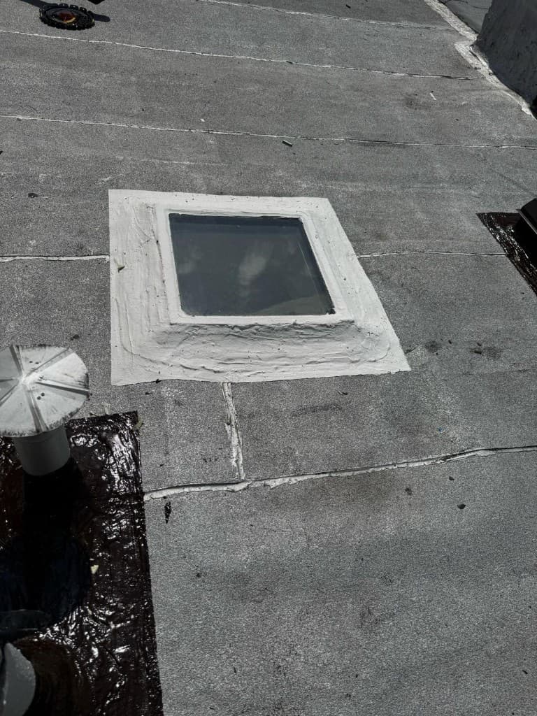 Skylight curb on a gray roof coated all around with thick gray mastic, with a vent pipe flashed in black cement at the left