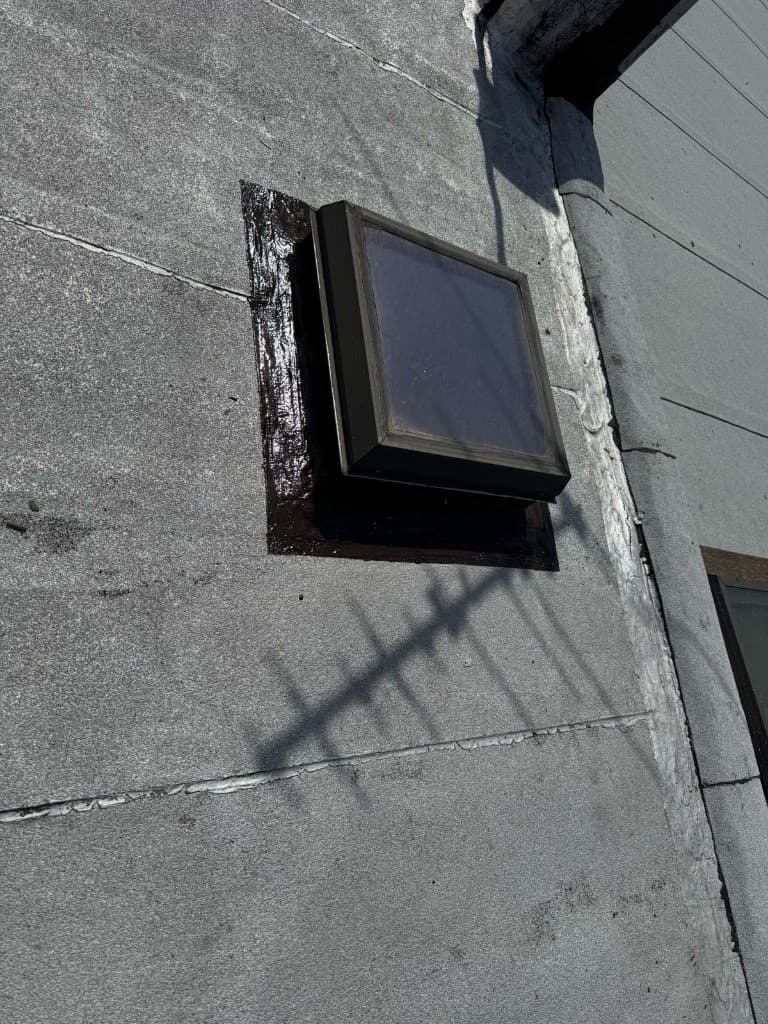 Small dark-framed skylight on a roof with a glossy black cement pad beside it and the base of the adjoining wall sealed with a white strip