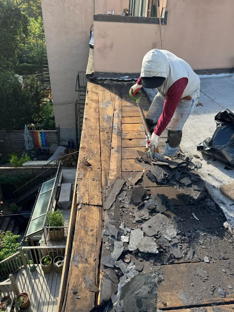 Roofer stripping old roofing off a wood plank deck along a parapet with a flat shovel, with debris and a trash bag beside him