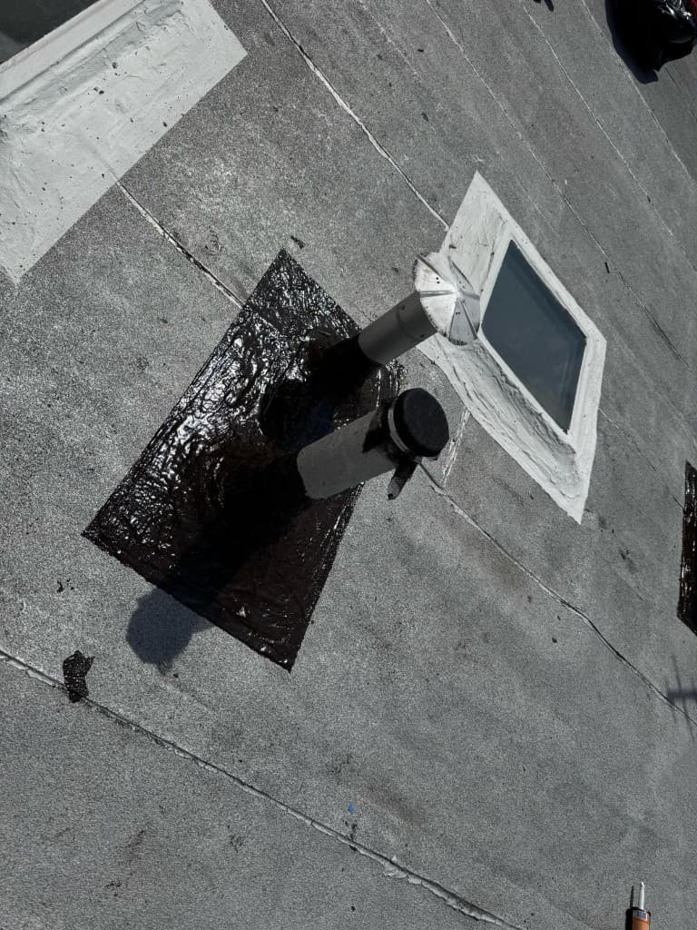 Two vent pipes with metal caps set in a glossy black cement flashing pad, with a skylight curb sealed in gray mastic behind them