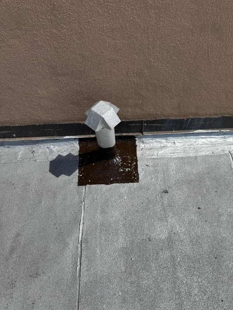 Vent pipe with an aluminum cap standing in a black cement flashing pad at the base of a tan stucco wall, with a black flashing strip and white finish along the wall