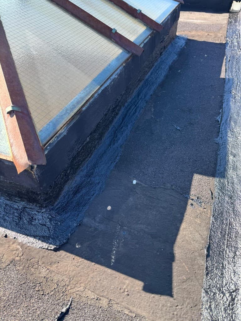 Close view of the base of a wire-glass skylight curb with black flashing material applied around it on a gray roof