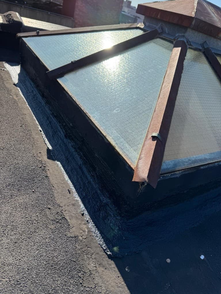 Wire-glass skylight with a rusty metal frame whose curb has been re-flashed with black flashing material on a gray roof