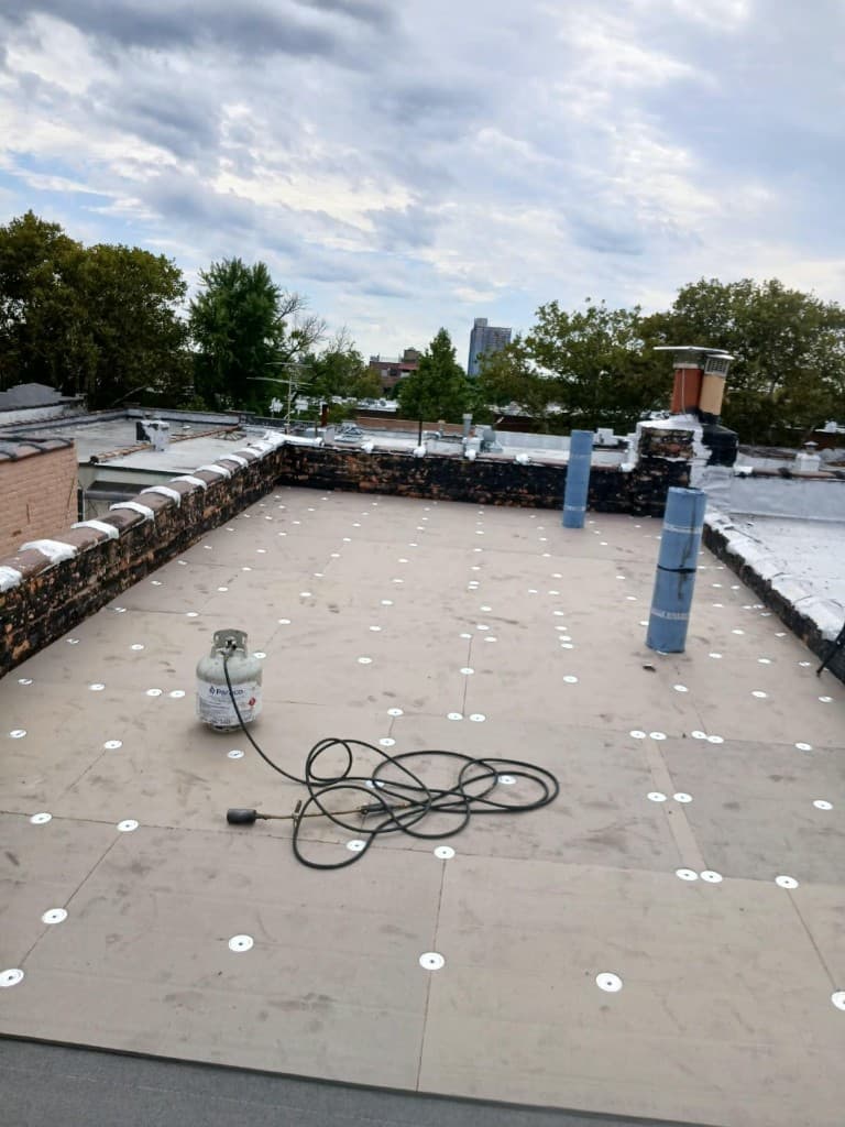 Flat roof covered with new boards fastened in a grid of round plates, with a propane torch and rolls of roofing material by the parapet