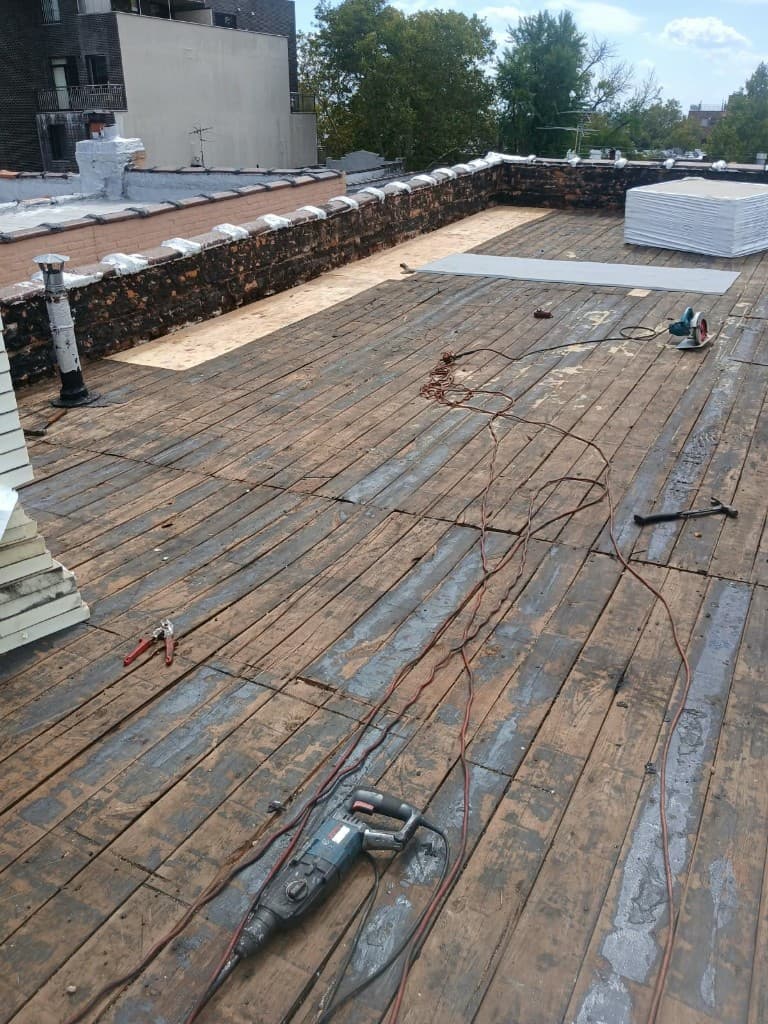Wood plank roof deck with new light-colored plywood laid along the brick parapet and materials stacked on the roof