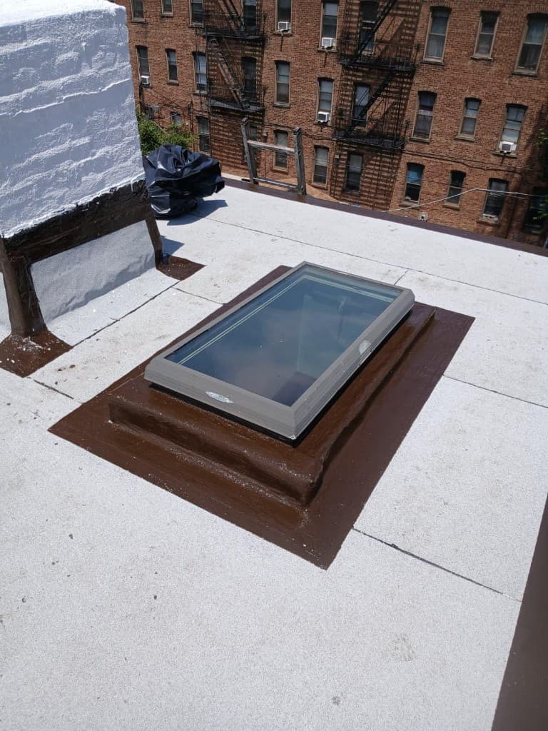 Skylight on a stepped curb coated in brown, with the coating carried out onto the gray cap sheet roof, next to a white-painted chimney