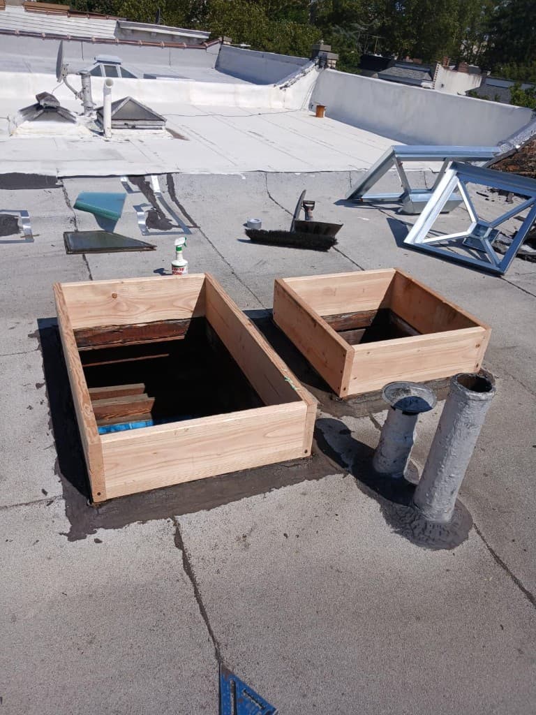Two new wood curbs framed around skylight openings on a flat roof, with new metal skylight frames laid out beside them and two vent pipes in flashing cement