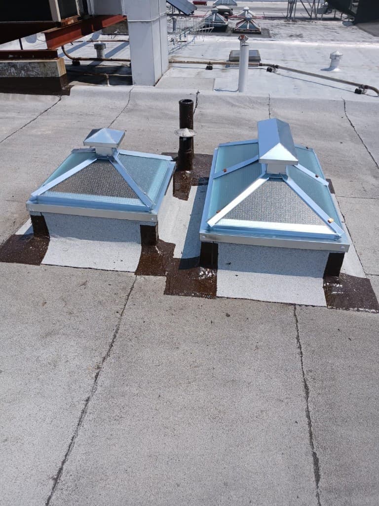 Two pyramid skylights with wired glass and metal frames on curbs wrapped in cap sheet, corners sealed in brown, with a vent pipe between them