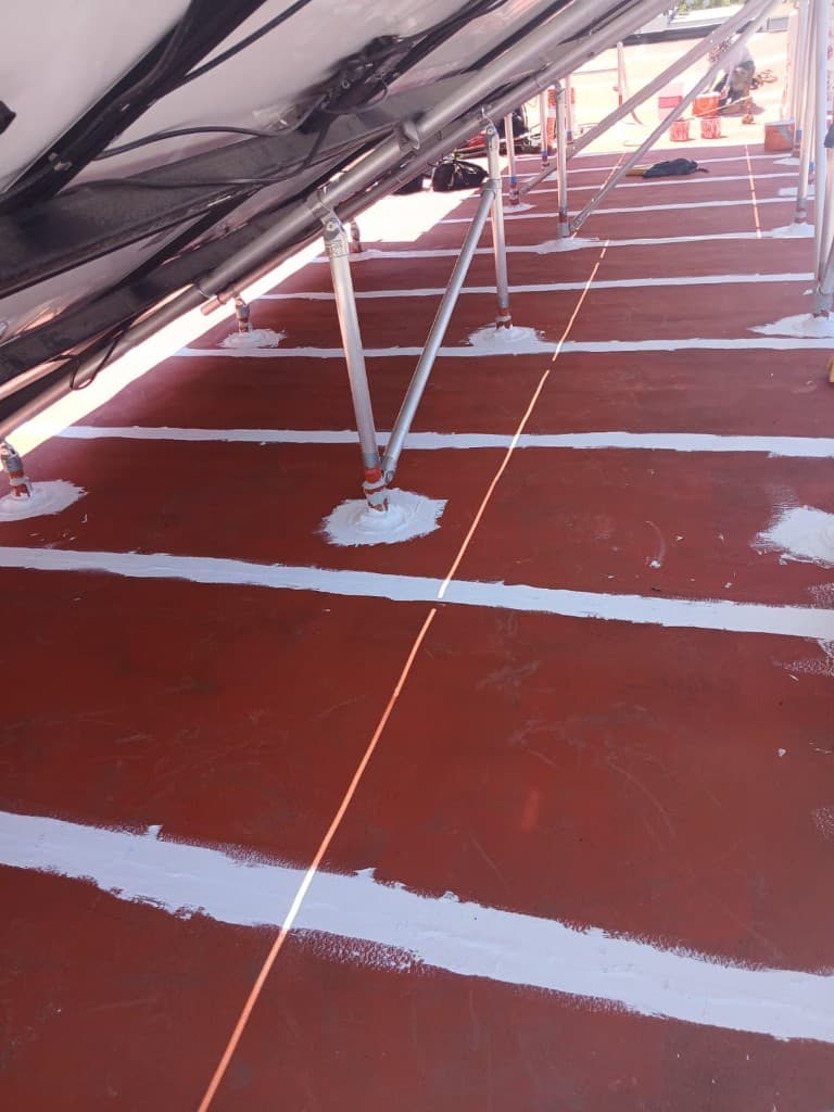 Red base coat with white-reinforced seams between rows of aluminum solar-array legs, each leg base sealed with a round pad of white coating