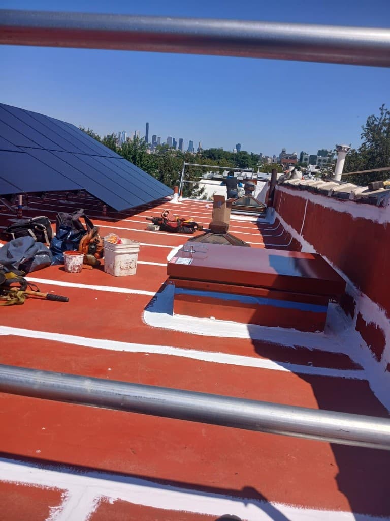 Roof with a red base coat and white-reinforced seams, solar panels on the left, skylights and a roof hatch edged in white, and tools staged on the roof
