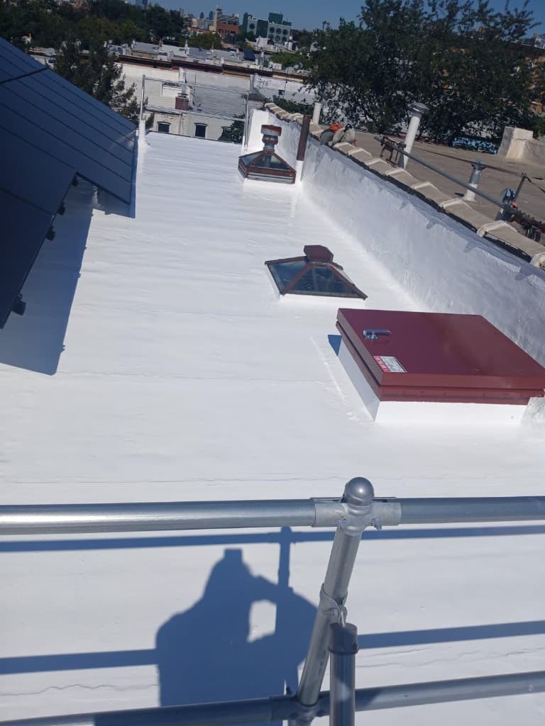 Finished white silicone coating across a flat roof with skylights, a red-covered roof hatch and vents sealed, beside a solar array