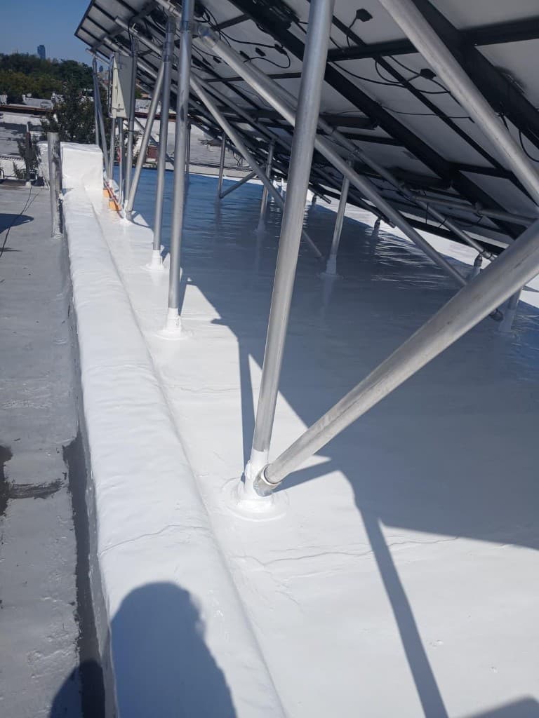 Finished white silicone coating around aluminum solar-array legs with each base sealed, and the roof-edge wall coated